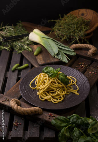 Italian spaghetti tasty appetizing classic pasta with basil on a dark plate on a wooden tray. In the background are greens, cucumbers, a knife and wooden utensils.