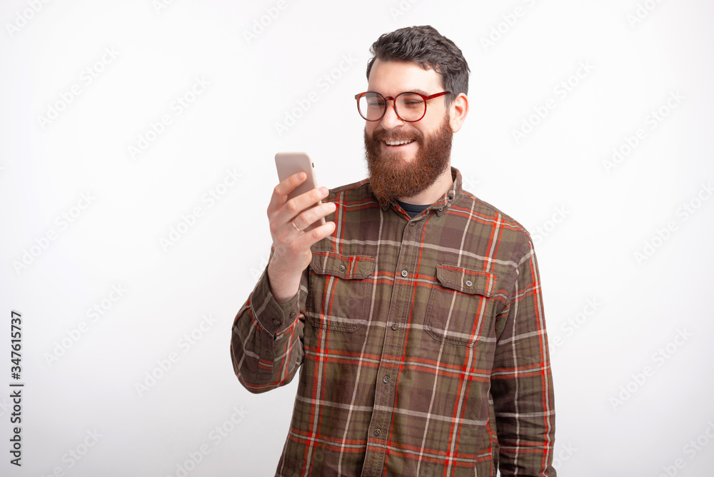 Handsome bearded man wearing glasses is looking at his phone, watching something on white background. Handsome bearded man wearing glasses is looking at his phone, watching something on white background.