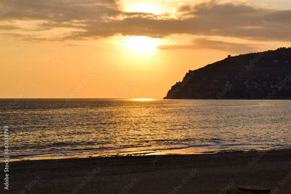 Golden sunset over the Mediterranean Sea. The setting sun shines through the clouds, and a golden path is visible above the ocean. Silhouetted mountains on the horizon.