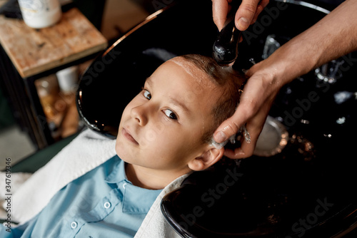 Hairwash. Cute little boy leaning on the sink in barbershop and looking at camera while male barber washing his hair photo