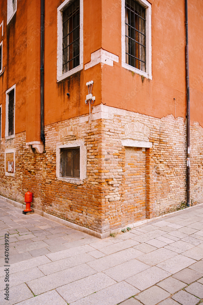 Corner of an orange red brick building. The second floor is plastered, the first floor is bare brick. Brick front door, metal bars on the windows. Red fire hydrant is standing against the wall.