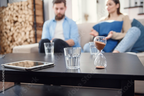 Sand clock at psychologist cabinet with unhappy annoyed caucasian couple having psychologist therapy session on background.