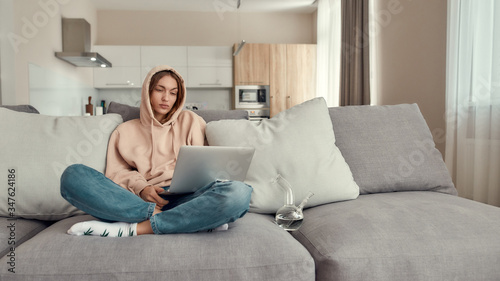 Too much relaxed. Young caucasian woman sitting on the couch at home, trying to use laptop after smoking marijuana from a bong or glass water pipe © Svitlana