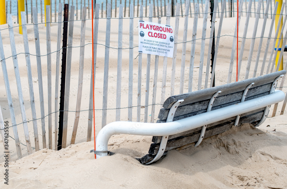 Sign saying playground at the beach is closed, and sand covers an empty park bench Sign saying playground at the beach is closed, and sand covers an empty park bench