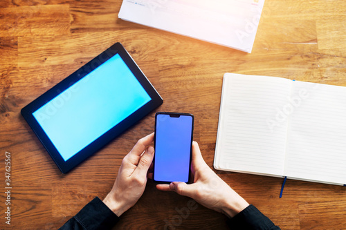 Flat lay. Male hands is using a phone with a blank screen, digital tablet, a notebook and working papers on the wooden table near. Top view, copy space