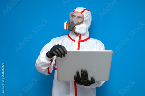 disinfection worker in protective suit and respirator checks coronavirus statistics on laptop, virologist on blue background