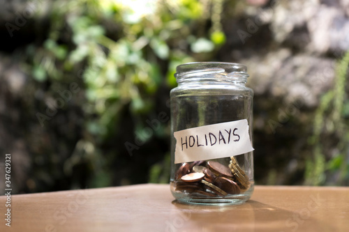 A glass jar with coins inside and a "vacaciones" tag on a wooden. Un bote de cristal con monedas dentro, tiene una etiqueta que pone vacaciones sobre una mesa de madera y fondo de plantas y piedras.