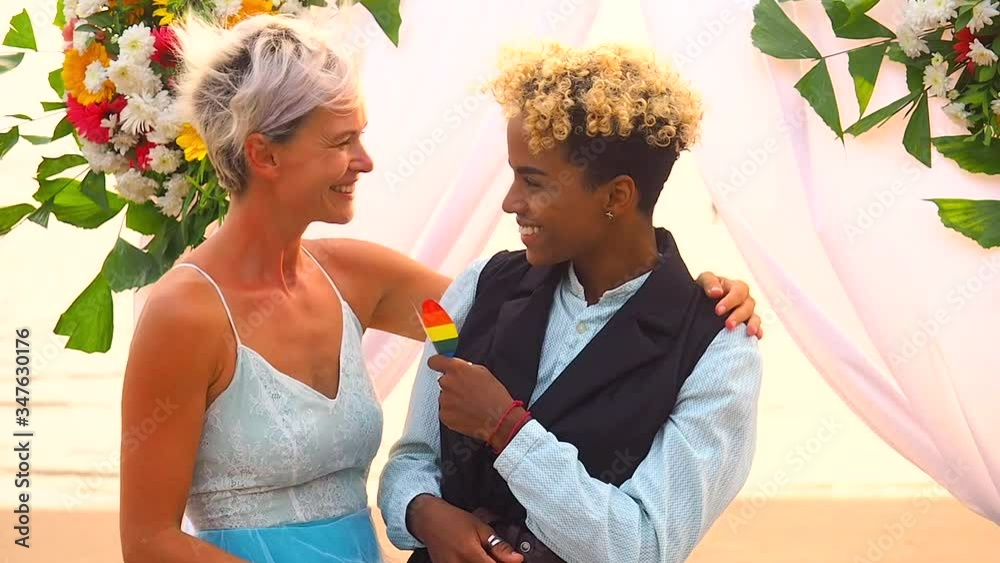 african american female groom in black suit and caucasian bride in dress in ceremony holding rainbow heart in hands on tropical beach under wedding flover arch