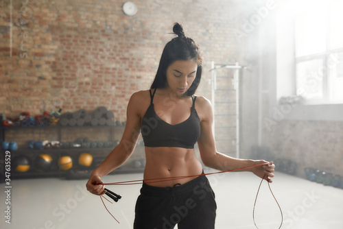 Move your body, shape your future. Sportive woman in black sportswear holding jump rope while having workout at industrial gym photo