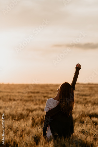 Young beautiful girl in white dress and jacket walking through the wheat orange field on a sunny summer day. Feel freedom with arm stretched to the sky. Nature in the country. Sunset on isolation