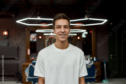 Perfect look. Portrait of happy young guy in white T-shirt looking at camera and smiling while visiting modern barbershop
