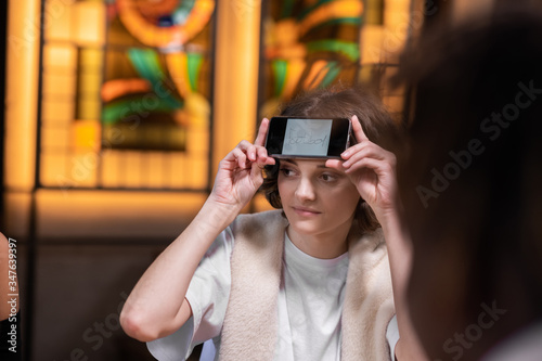 A pretty girl with her smartphone on her forehead sitting at the table with friends and trying to guess a word written on her display, close up picture