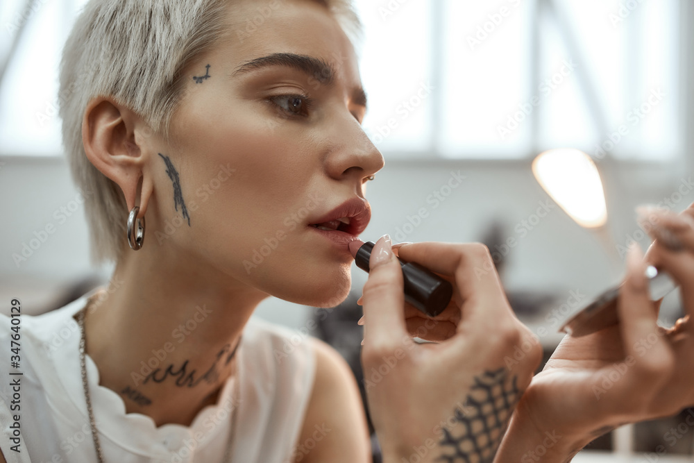 Looking perfect. Portrait of young and stylish blonde tattooed business woman applying lipstick while sitting in the modern coworking space