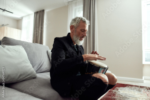 Let the Music Speak. Bearded middle-aged man, artist or composer writing song, playing rhytm with djembe drum, sitting on the couch at home