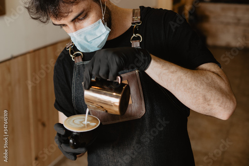 camarero barista haciendo un dibujo latte art en una taza negra de café, lleva mascarilla y guantes covid-19 photo