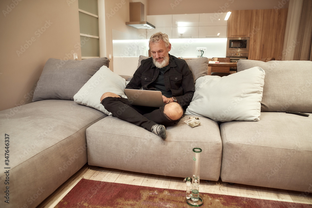 Working indoors. Bearded middle-aged man using laptop while sitting on the couch. Bong or glass water pipe, buds in plastic bag and lighter near him. Cannabis and weed legalization concept Working indoors. Bearded middle-aged man using laptop while sitting on the couch. Bong or glass water pipe, buds in plastic bag and lighter near him. Cannabis and weed legalization concept