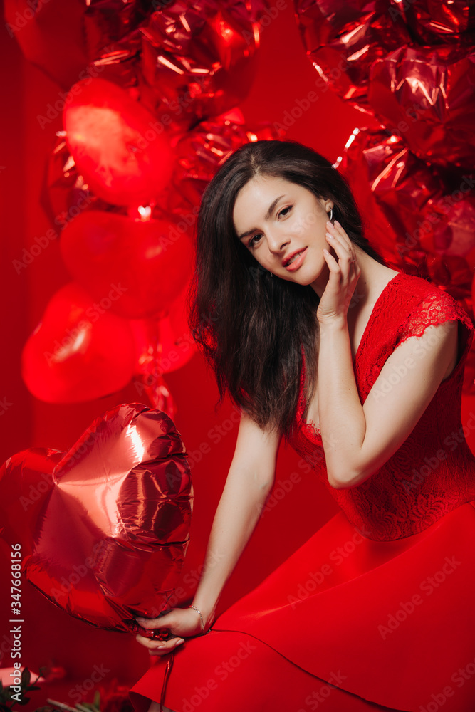 Woman with Hearts Balloons on a red background. Beauty Girl on Valentine's Day. Model in a stylish red dress.