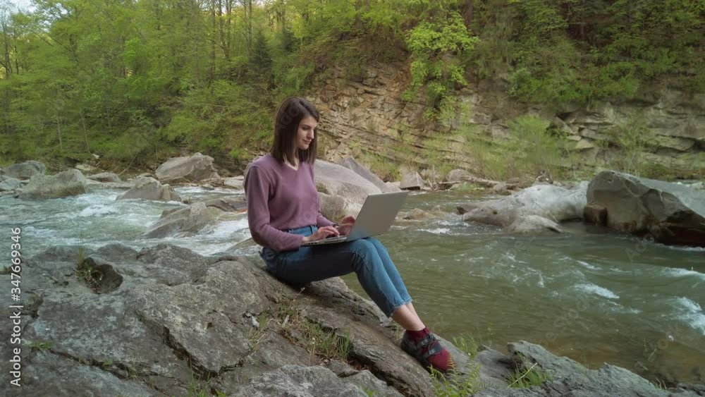 Female IT specialist with a laptop works outdoors near a mountain stream. Sitting on a rock. Beautiful nature around. Female IT specialist with a laptop works outdoors near a mountain stream. Sitting on a rock. Beautiful nature around.