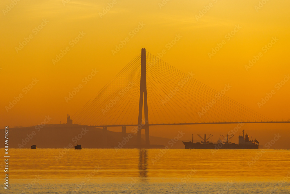Obraz premium Dawn in the sea city of Vladivostok. A large commercial cargo ship stands on a roadstead against a cable-stayed Russian bridge in Vladivostok during a bright golden dawn