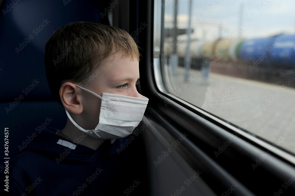 sad blond boy preschooler in a white medical mask sits on a train and looks out the window at a train passing by. Wear a mask. Taking care of yourself during a pandemic. Travel ban