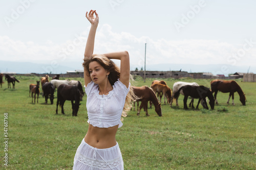 Beautiful girl in a white dress on a background of grazing horses