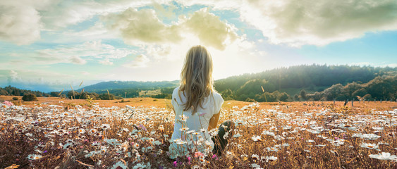 Woman sitting on the chamomiles field and looking at the cloudy sky at sunset. Banner edition..