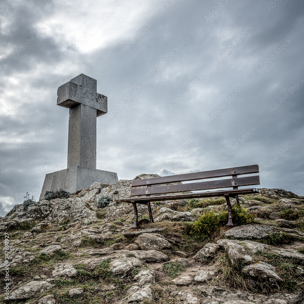 Square format photography of an old weathered stone calvary near a bench sealed at the top of a rocky hill