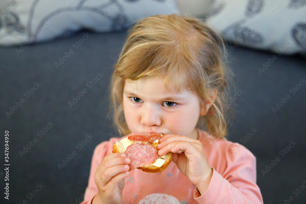 Adorable little toddler girl eating fresh prepared salami sandwith in kitchen. Happy child eats healthy food for lunch or dinner. Baby learning. Home, nursery, playschool or daycare