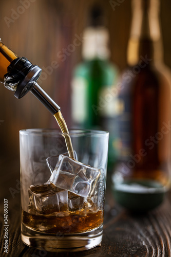 In the bar, whiskey is poured into a glass with ice, in the background there are bottles on a wooden table of the bar counter, shallow depth of field, selective focus. The concept of alcoholic drinks