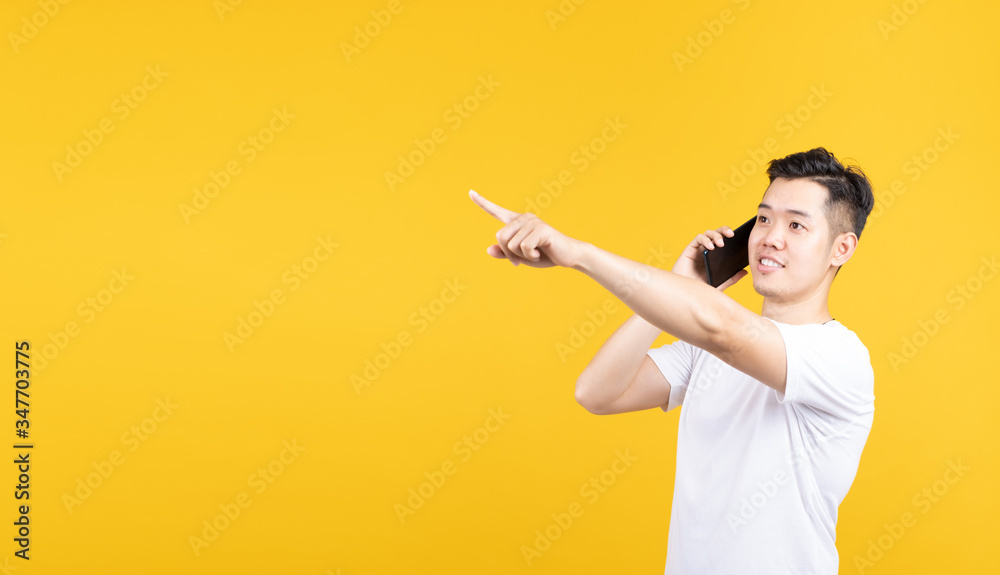 Portrait happy young asian man talking with mobile phone smiling pointing side for advertising banner with copy space wearing white t-shirt on yellow background isolated studio shot.