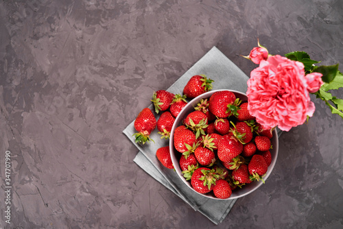 Ripe strawberries in ceramic bowl on linen table napkin and pink rose in vase on concrete background, copy space. Healthy food concept, still life. Top view, flat lay