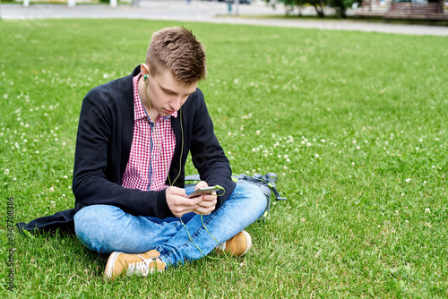 Portrait of happy young student sitting on green grass lawn in park, listening to music with earphones and using mobile smartphone outdoors, copy space