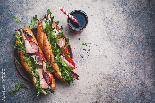 Two fresh baguette sandwiches with meat, tomato, cucumber and arugula on gray background, top view.