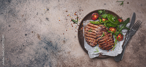 Grilled beef steaks with arugula and tomato salad on wooden plate, dark background, copy space, top view.