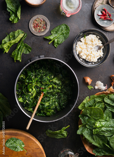 Spinach cooking preparation. Stewed spinach in black cooking pot with spoon on rustic background with ingredients and kitchen utensils . Top view. Home cuisine. Healthy food nutrition
