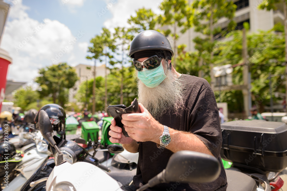 Mature bearded hipster man with sunglasses and mask using phone while sitting on motorcycle