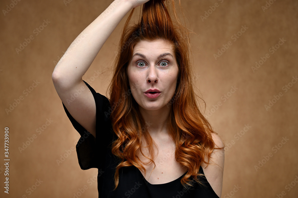 Naklejka premium Studio large photo portrait of Caucasian young actress woman with long red hair on a beige background. Model poses with a smile and grimace.