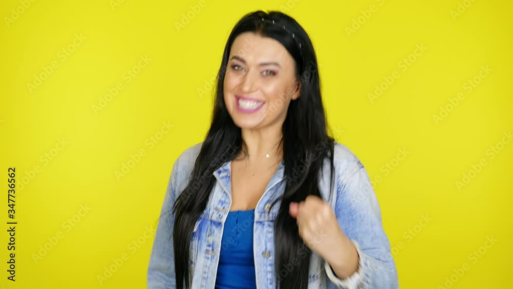 Happy woman with black hair in a denim jacket rejoices in victory, waves her hands and says yes on a yellow background with copy space. Place for text or product. 4K footage Happy woman with black hair in a denim jacket rejoices in victory, waves her hands and says yes on a yellow background with copy space. Place for text or product. 4K footage