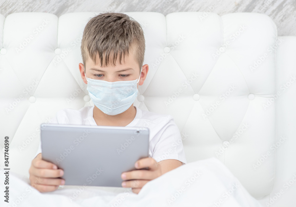 Sick boy wearing medical protective mask uses tablet computer on the bed at home. Quarantine and coronavirus epidemic concept. Empty space for text Sick boy wearing medical protective mask uses tablet computer on the bed at home. Quarantine and coronavirus epidemic concept. Empty space for text