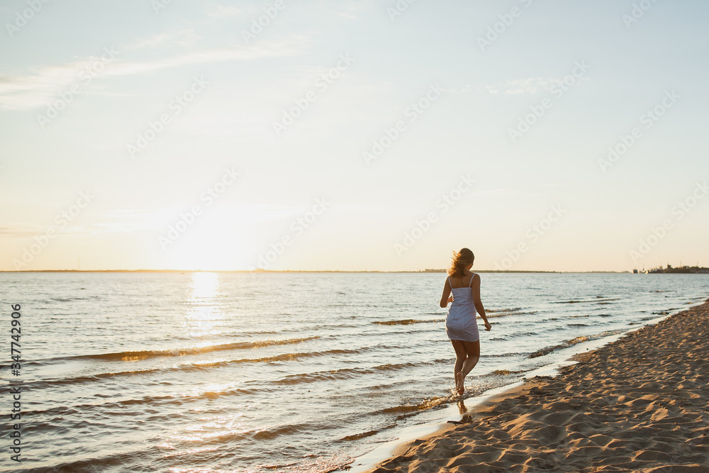 Playful tanned woman running on the beach on the sunset . Cheerful young woman with perfect shape in sexy dress enjoying summer sunset. Dating, carefree summer concept.