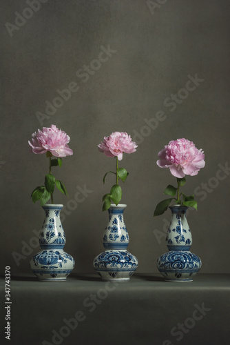 Three delft blue vases with pink peonies on a table in a grey room.