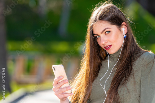 young smiling woman sit on bench in park outdoors listening music with headphones using mobile phone on a sunny and windy day