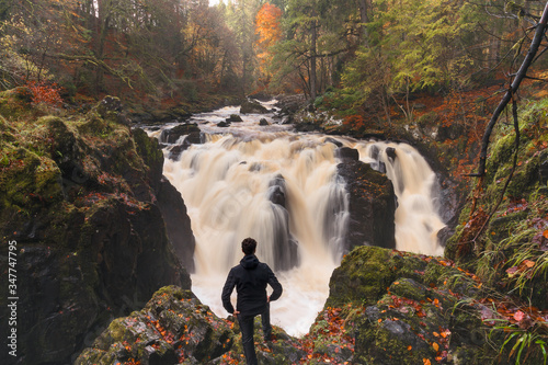 A man standing on a cliff covered with red leaves,  watches with satisfaction a beautiful waterfall of the Hermitage, Scotland in Autumn.  photo