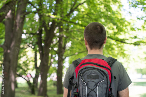 A young male tourist is walking in the park with a gray-red backpack. Back view. Nature travel concept with place for text.