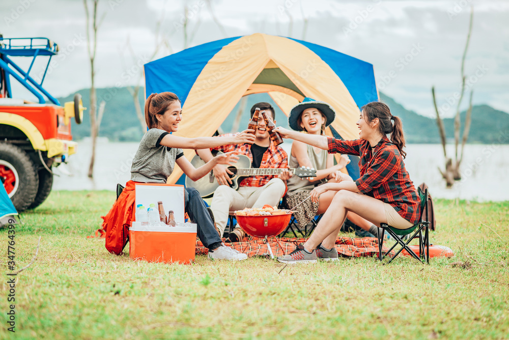 Two Asian women toasting bottles of beer to each other to celebrate a good time with a group of friends while traveling on a camping tent on holiday. - obrazy, fototapety, plakaty 