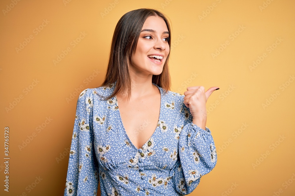 Young beautiful brunette woman wearing casual floral t-shirt standing over yellow background smiling with happy face looking and pointing to the side with thumb up. Young beautiful brunette woman wearing casual floral t-shirt standing over yellow background smiling with happy face looking and pointing to the side with thumb up.