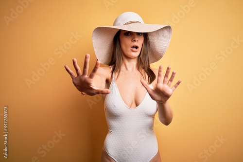 Young beautiful brunette woman on vacation wearing swimsuit and summer hat doing stop gesture with hands palms, angry and frustration expression