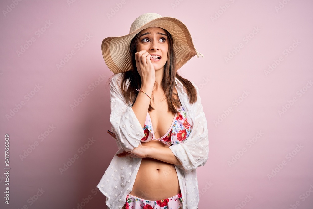 Young beautiful woman on vacation wearing bikini and summer hat over pink background looking stressed and nervous with hands on mouth biting nails. Anxiety problem. Young beautiful woman on vacation wearing bikini and summer hat over pink background looking stressed and nervous with hands on mouth biting nails. Anxiety problem.