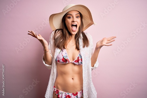 Young beautiful woman on vacation wearing bikini and summer hat over pink background celebrating crazy and amazed for success with arms raised and open eyes screaming excited. Winner concept