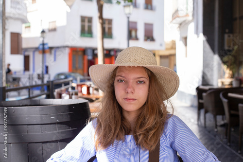 A cute girl of Caucasian ethnicity with long hair in a straw hat is resting in a street cafe on a summer day. © Ирина Селина